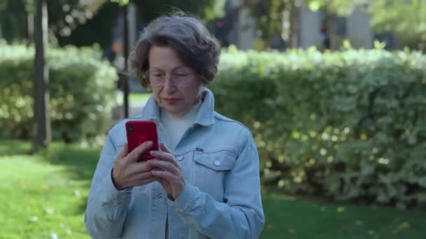 高齢者ポジティブな女性は 晴れた秋や春の日に市立公園の屋外に立って散歩に出かけます 笑顔の女性はスマートフォンを使用し ソーシャルで面白いビデオを見ます — ストック動画