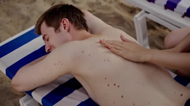 ガールフレンドがスキンクリームを塗ったときに日焼けに横たわっている若い男 女性は休暇中にビーチで眠っている日焼け止めのリラックスしたボーイフレンドを適用します 海辺の近くでカップルを冷やしている マッサージをする少女 嘘をつく — ストック動画