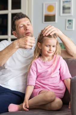 Genç bir baba ve küçük bir kızdan gelen cana yakın bir aile odadaki kanepenin yanında oturuyor. Şefkatli bir baba saçlarını saç tokalarıyla süsler. Mutlu çocukluk ve babalık kavramı. Kapat.
