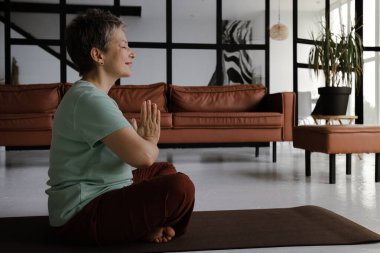 Orta yaşlı bir kadın büyük salonda yoga yapıyor. Gülümseyen bir kadın, elleri ve gözleri kapalı, göğsünde bir araya gelmiş, nilüfer pozisyonundaki bir paspasın üzerinde yan yan oturuyor. Konsept: