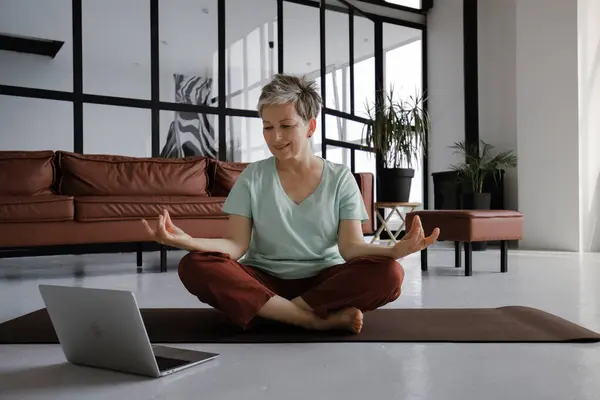 A middle aged lady is practicing yoga at home. A woman with slight smile on face sits on a mat on the floor in the lotus position, uses a laptop to watching relaxing videos and listening meditative
