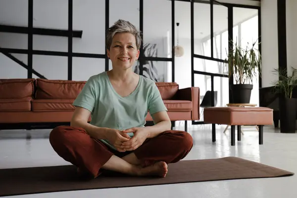A middle aged lady rests after practicing yoga in the large hall of the house. A smiling woman sits on a mat on the floor in the lotus position in a happy mood after pleasant workout. Looks at the