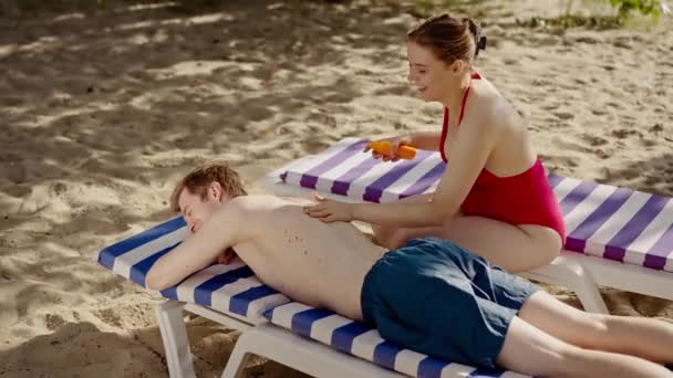 ビーチでリラックスする若いカップル 赤いビキニのガールフレンドは 彼女のボーイフレンドスパフクリームを適用します 屋外でのリゾート休暇に日焼け止めを使用した男女の観光客 女性は夫に日焼け止めをこすります — ストック動画