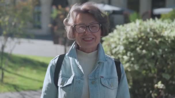屋外の公園に立っている光り輝く笑顔の高齢女性 市内中心部のデニムシャツの美しい老婦人の肖像画 美しい旅行者女性年金は街の外で陽気です 高品質4Kについて — ストック動画