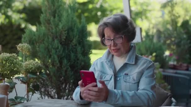 屋外テラスに座っているときにスマートフォンを使用して大人の幸せな灰色の髪の女性 カフェで携帯をタップするシニア女性 老婦人は食事メニューを見つけるためにカフェテリアでQrコードをスキャンしました 高品質の4K映像 — ストック動画