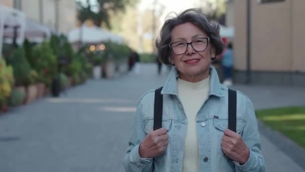 アクティブな観光客 シティパークの背景に背後にあるリュックサックを持つ高齢女性 陽気な魅力的な年金女性は 新しい都市で幸せです 美しい女性の古い旅行者のトゥーティーな笑顔 高品質4Kについて — ストック動画
