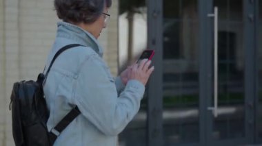 Harita uygulaması kullanarak akıllı telefonlu yaşlı bir gezginin yan yürüyüşü. Yeni şehirde doğru yolu bulmak için telefon arayan yaşlı bir turist kadın. Sırt çantasıyla gezen emekli cep telefonuyla gezen
