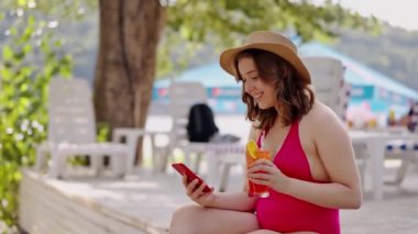 Vücut pozitifliği olan bir kız tatilde sosyal medyada geziniyor. Kırmızı bikinili kadın yaz tatilini dışarıda havuz başında geçiriyor. Cep telefonu kullanarak mayo giymiş neşeli bir bayan içerken blog yazar.
