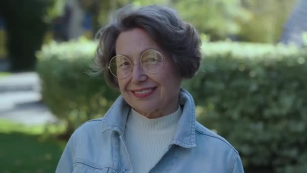 ミッドショット 美しくて幸せな祖母は屋外でスタイリッシュな眼鏡でデニムシャツを着ていました 都会公園の可愛い女性年金受給者のポジティブな笑顔 外で微笑んでいるランダムな老婦人 — ストック動画
