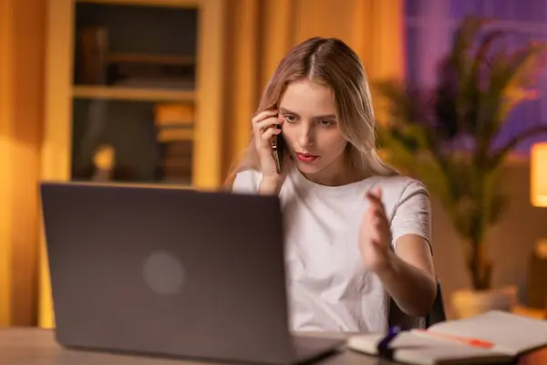 A serious girl student sits at a table in apartment, uses a laptop to search for educational materials and smartphone to communicate with fellow student. A young lady teacher remotely indicates errors