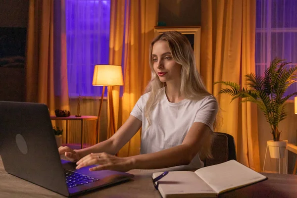 一个女生坐在公寓的桌子旁 使用笔记本电脑 打字作业或论文 一位未来的女毕业生为即将到来的考试作准备 直到深夜 一个可爱的金发女人 — 图库照片