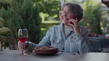 Mutlu yaşlılık kavramı. Park 'taki bir restoranda öğle yemeği sırasında telefon eden emekli bir kadın. Gülümseyen yaşlı bayan akıllı telefondan konuşuyor ve kafede şarap içiyor. Yüksek kalite 4k görüntü