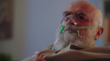 Hasta, yaşlı bir adam evde tek başına tedavi eder, nefes almak için oksijen maskesi kullanır ve ısınmak için ekoseyi omuzlarına sarar. Hasta bir insan koronavirüsten ya da bronşiyal astımdan muzdariptir.
