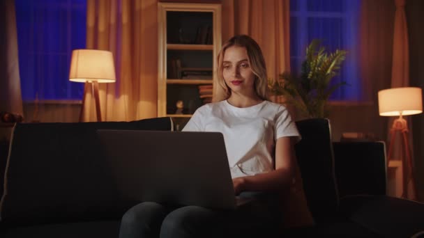 ブロンドの女の子がソファーに座り 仕事の日の後に夜の余暇を過ごし ラップトップを使って電子ブックを読んだり 友人と対応したりします 若いビジネスマンがオンラインストアの運営をコントロール — ストック動画
