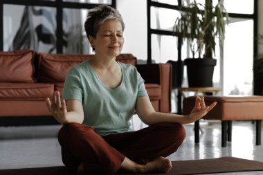 Orta yaşlı bir bayan evde yoga yapıyor. Olumlu bir ruh hali içinde, gözleri kapalı ve hafifçe gülümseyen bir kadın lotus pozisyonundaki bir paspasın üzerinde rahatça oturur. Manevi gelişim kavramı