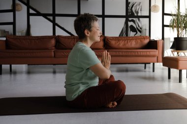 Orta yaşlı bir kadın büyük salonda yoga yapıyor. Yoğunlaşmış bir kadın, gözleri ve avuçları kapalı, göğsüne katlanarak, nilüfer pozisyonundaki bir paspasın üzerinde yan yatar. Konsept
