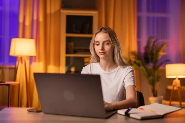 一个专心致志的女生坐在公寓的桌子旁 用笔记本电脑 打字或写论文 一位严肃的未来女毕业生将为即将到来的国家考试做准备 直到深夜 — 图库照片