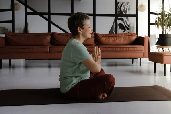 A middle aged lady practices yoga in the large hall. A concentrated woman with closed eyes and palms, folded together on chest, sits sideways on a mat on the floor in the lotus position. The concept