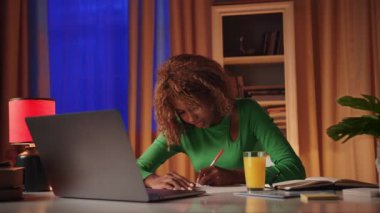 Genç bir Afrikalı kız öğrenci sınava hazırlanmak için gerekli bilgileri aramak için bir dizüstü bilgisayar kullanıyor ve sol elini kullanarak bir deftere yazıyor. Bir bayan yabancı dil öğreniyor, monitöre bakıyor.