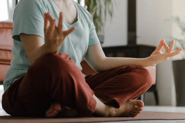 A middle aged lady is practicing yoga at home. A woman without a face sits relaxed sideways on a mat on the floor in the lotus position. The concept of spiritual development through physical exercise