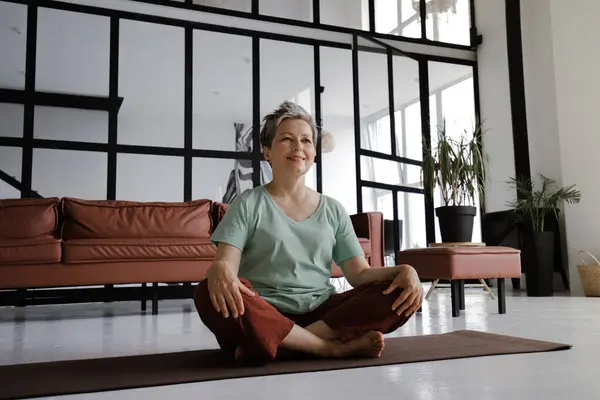 Orta yaşlı bir bayan, evin büyük salonunda yoga yaptıktan sonra dinleniyor. Gülümseyen bir kadın, hoş bir egzersizden sonra lotus pozisyonunda yerde mutlu bir şekilde oturur. Şuna bak.
