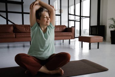 Orta yaşlı bir kadın evde yoga yapıyor. Mutluluktan gözleri kapalı gülümseyen bir kadın lotus pozisyonunda yerde bir paspasın üzerine oturur ve kollarını başının üstüne uzatır. Vücudun uyumu kavramı