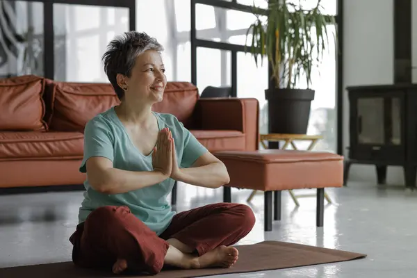 A middle aged lady is practicing yoga at home. A woman with closed eyes and palms, folded together on chest, sits on a mat on the floor in the lotus position. The concept of spiritual development