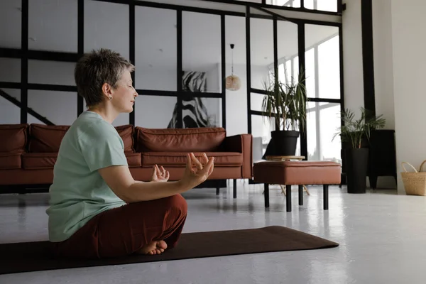 A middle aged lady is practicing yoga in the large hall of the house. A woman with closed eyes sits relaxed sideways on a mat on the floor in the lotus position. The concept of spiritual development