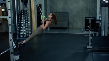 Spor salonundaki genç bir kız sporcu esnemek için spor kemerlerini bağlar. Bir erkek fitness eğitmeni onu destekliyor. Kol gücü ve göğsün güzelliği için egzersiz. Yüksek kalite 4k