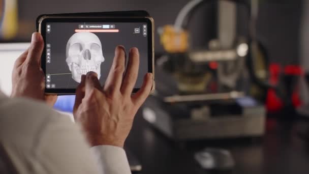 男性のグラフィックデザイナーが手に持つタブレットを使用して 歯を持つ人間の頭蓋骨の3Dモデルを表示します 3Dプリンターの移植のためのバイオプリンティング臓器の概念 医学の未来について クローズアップ — ストック動画