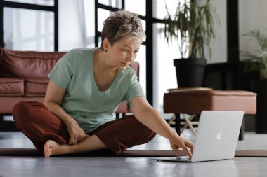 Orta yaşlı bir bayan büyük salonda yoga eğitimi aldıktan sonra rahatlıyor. Yoğunlaşmış bakışlı bir kadın, spor programı oluşturmak için bilgisayarlı bir akıllı telefon kullanır, en uygun zamanı hesaplar.