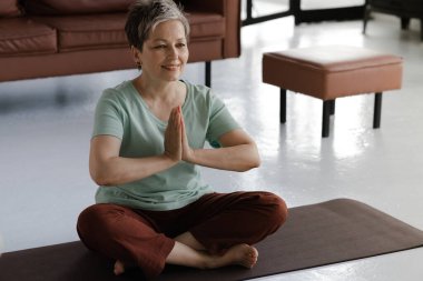Orta yaşlı bir kadın evde yoga yapıyor. Gözleri kapalı bir kadın lotus pozisyonunda yerde bir hasırın üzerine oturur ve avuçlarını kaldırıp namaste hareketi gösterir. Kapatın. Yüksek kalite