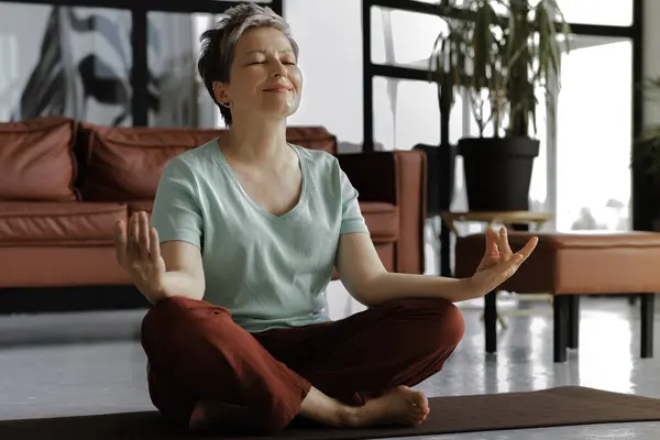 A middle aged lady is practicing yoga at home. A woman in positive mood with closed eyes and slight smile sits relaxed on a mat on the floor in the lotus position. The concept of spiritual development