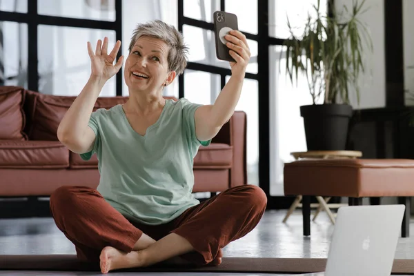 A middle aged lady make video about yoga in the large hall of the house. A smiling woman sits on a mat on the floor in the lotus position, has a break time from training, uses a smartphone for video