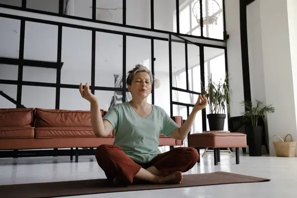 A middle aged lady is practicing yoga at home. A woman with closed eyes and smile on a face sits relaxed on a mat on the floor in the lotus position. The concept of spiritual development through
