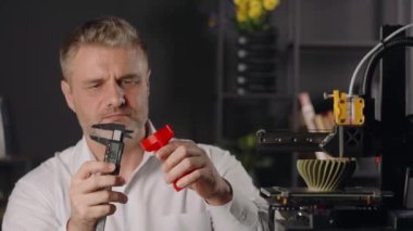 Orta yaşlı bir grafik tasarımcısı dijital kalibreler kullanır ve kırmızı plastik huninin parametrelerini ölçer. Otomatik özerk süreçte çalışan ve haki rengi üreten 3D yazıcı makinesi