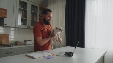 Mutlu, genç, sakallı, sakallı bir adam, bir evcil hayvan arkadaşıyla mutfakta duruyor, beyaz bir köpek, chihuahua cinsi, okşuyor, kollarına alıyor ve sevgiyle tutuyor. Girişimci dizüstü bilgisayar kullanır