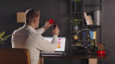 Bir adam grafik tasarımcısı masanın yanında oturur, kalibreler kullanır ve kırmızı plastik huninin parametrelerini ölçer. Dizüstü bilgisayarın ön planında geometrik formda vazo çizimi. Bir 3d yazıcı makinesi