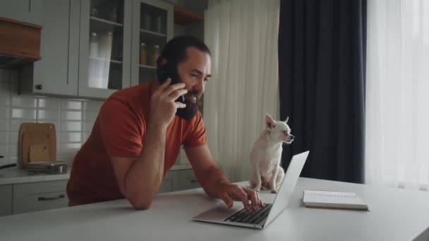一个留着胡子的年轻人站在厨房的一边 陪着他的一个宠物朋友 一只养着奇瓦瓦的白狗 抚摸着它 一个面带微笑的创业者在家里远程工作 用笔记本电脑 — 图库视频影像