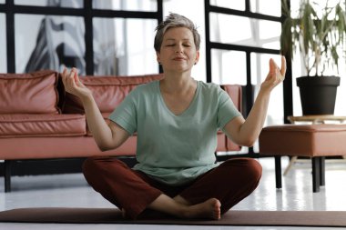 Orta yaşlı bir bayan evde yoga yapıyor. Kapalı gözlü ve yüzünde gülümseme olan bir kadın lotus pozisyonundaki bir paspasın üzerinde rahatça oturur. Ruhsal gelişim kavramı