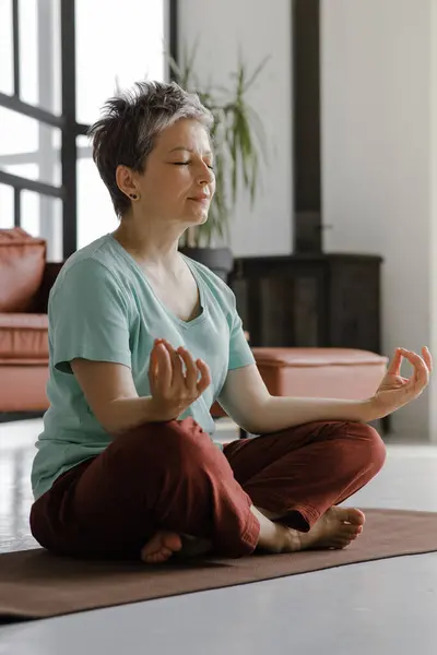 A middle aged lady is practicing yoga at home. A woman with closed eyes sits relaxed sideways on a mat on the floor in the lotus position. The concept of spiritual development through physical
