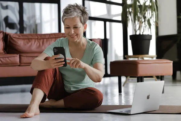 A middle aged lady rests after training yoga in the large hall. A brightly smiling woman sits on a mat on the floor, uses a smartphone and laptop for communication, searching and reading information
