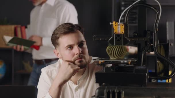 興味を持った集中的な若い男の機械工学者は 3Dプリンターの作業プロセスを観察します デジタル機械はカーキ色の幾何学的な形を作ります 中年男性の科学者が見て — ストック動画