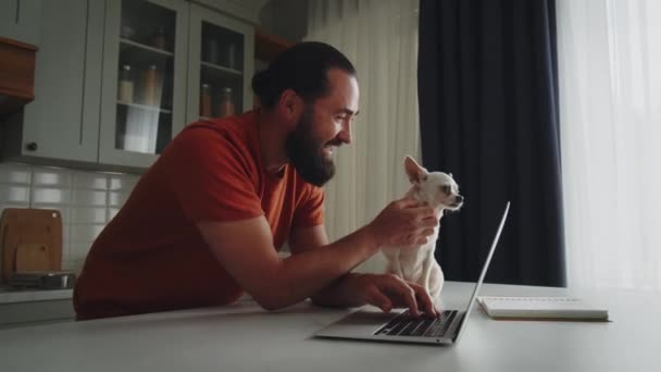 一个快乐的 笑容满面的年轻男人和他的宠物朋友 一只养着奇瓦瓦的白狗侧身站在厨房里 爱抚着它 亲切地抱着它 创业者用笔记本电脑打字 — 图库视频影像