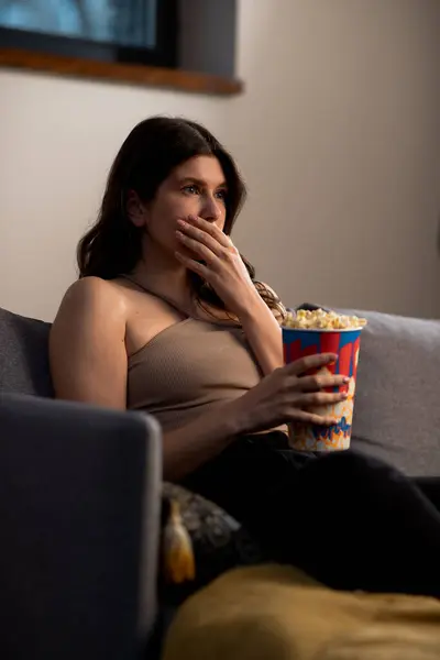 Surprised young woman is watching TV with cup of popcorn. Movie time at home. Girl sitting on sofa. High quality photo
