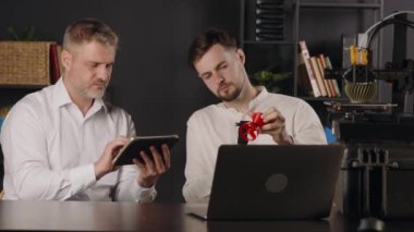 Genç bir makine mühendisi dijital kalibreler kullanır ve kırmızı plastik huninin parametrelerini ölçer. Orta yaşlı bir grafik tasarımcısı sonuçları tablete kaydeder. 3D yazıcı haki rengi yapar