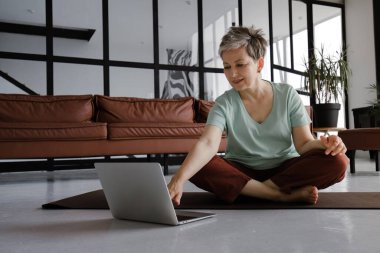 Orta yaşlı bir bayan evde yoga yapıyor. Gülümseyen bir kadın Lotus pozisyonunda yerde oturuyor, antrenmana ara veriyor, dizüstü bilgisayar kullanıyor ve gülüyor, dikkat dağıtıcı komik şeyler izliyor.