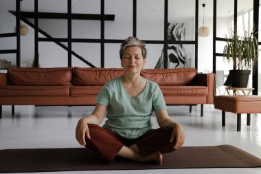 Orta yaşlı bir bayan, evin büyük salonunda yoga yaptıktan sonra dinleniyor. Gülümseyen bir kadın, hoş bir egzersizden sonra lotus pozisyonunda yerde mutlu bir şekilde oturur. Şuna bak.