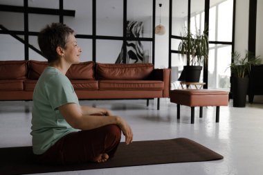 Orta yaşlı bir kadın büyük salonda yoga yapıyor. Yoğunlaşmış bir kadın, gözleri ve avuçları kapalı, göğsüne katlanarak, nilüfer pozisyonundaki bir paspasın üzerinde yan yatar. Konsept