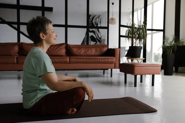 A middle aged lady practices yoga in the large hall. A concentrated woman with closed eyes and palms, folded together on chest, sits sideways on a mat on the floor in the lotus position. The concept